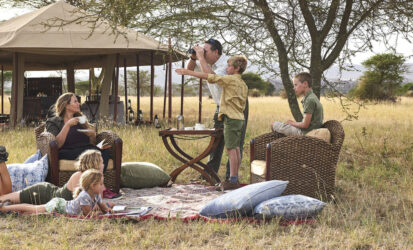 Family relaxing on a picnic blanket outside a safari tent, with a child pointing at wildlife