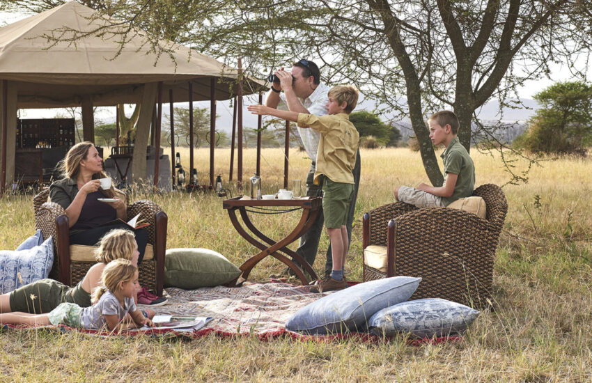 Family relaxing on a picnic blanket outside a safari tent, with a child pointing at wildlife